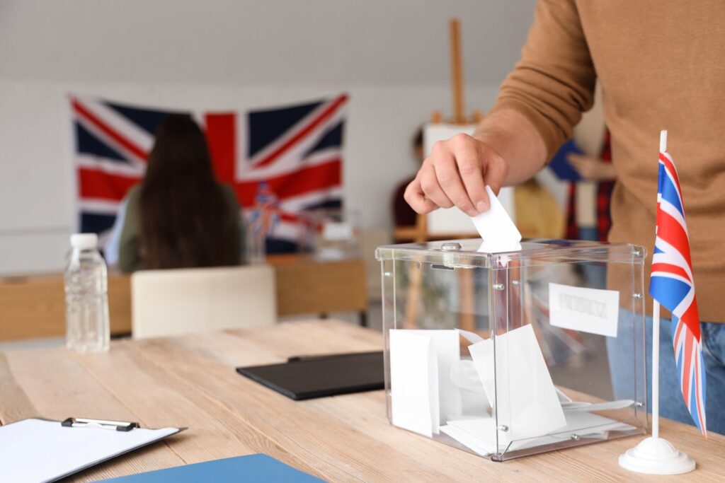 Voter placing a ballot paper into a clear ballot box, with Union flags in the background