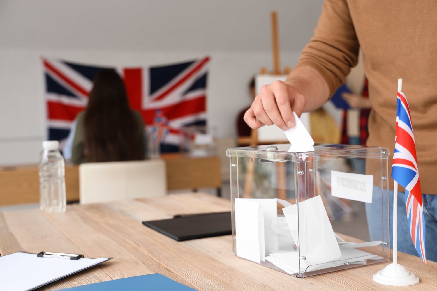 Voter placing a ballot paper into a clear ballot box, with Union flags in the background
