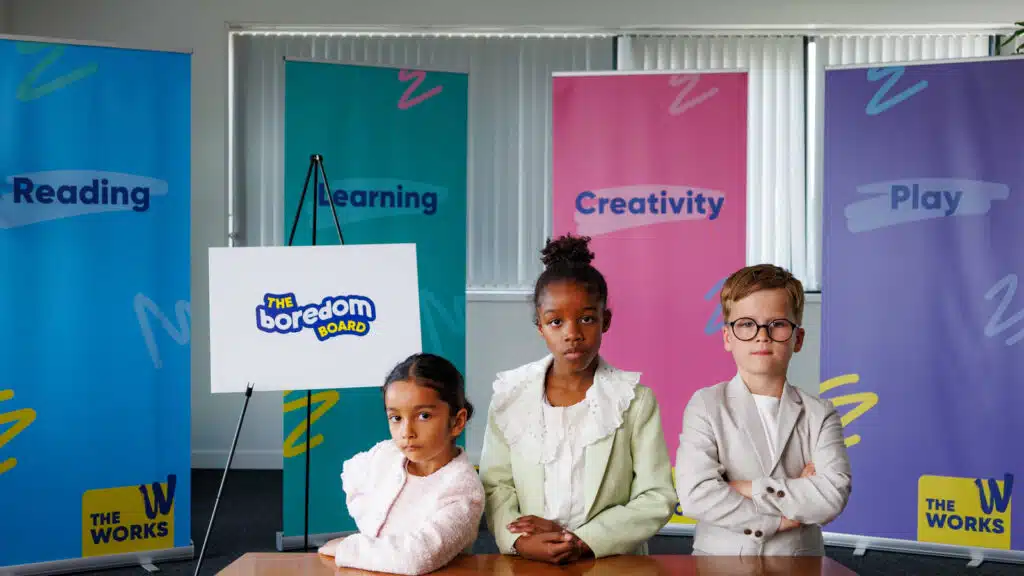 Three children at a desk in front of The Works 'Boredom Board' banners