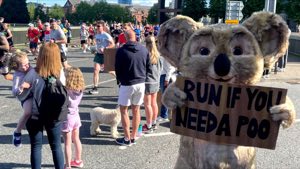 Koala mascot holding a sign at a running event as runners pass