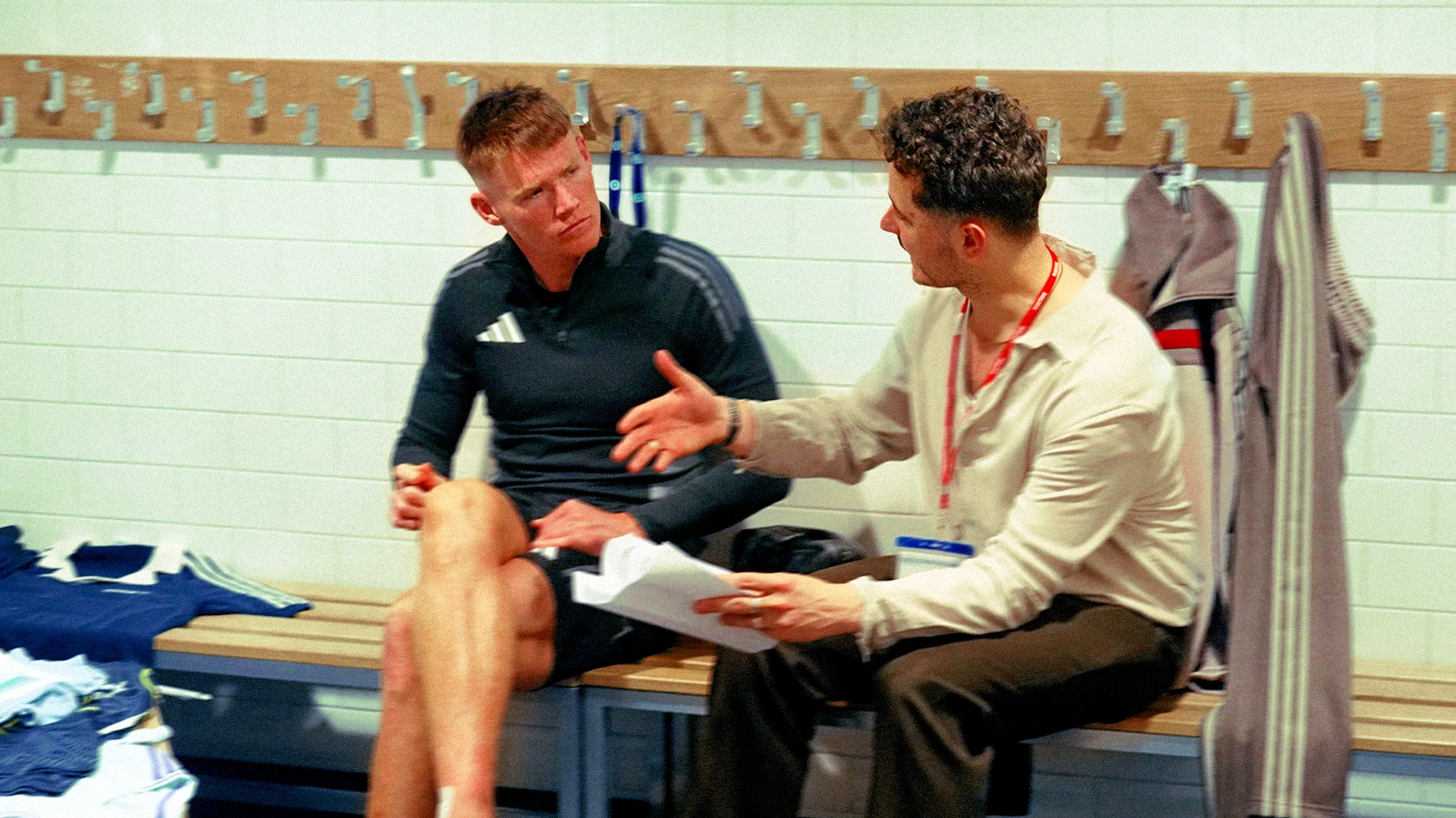 Two men chatting in a football changing room, one in training kit